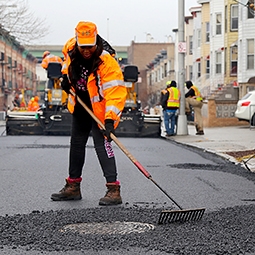 A NYC DOT construction worker in a neon orange jacket laying down new asphalt on a street